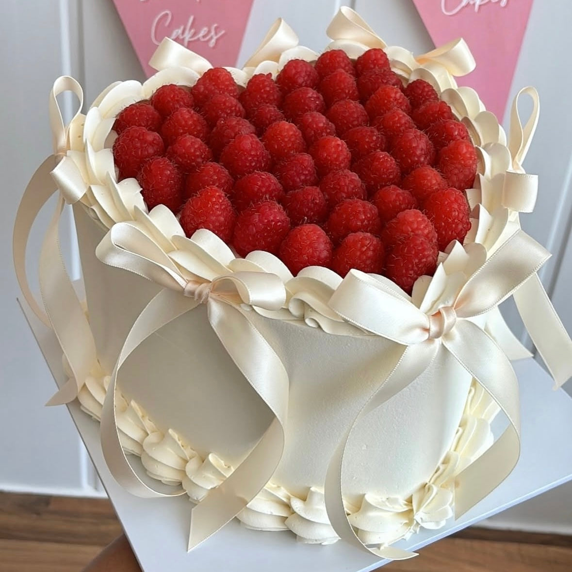 Elegant heart cake featuring piped frosting borders, ivory satin bows, and a topping of fresh, vibrant raspberries.