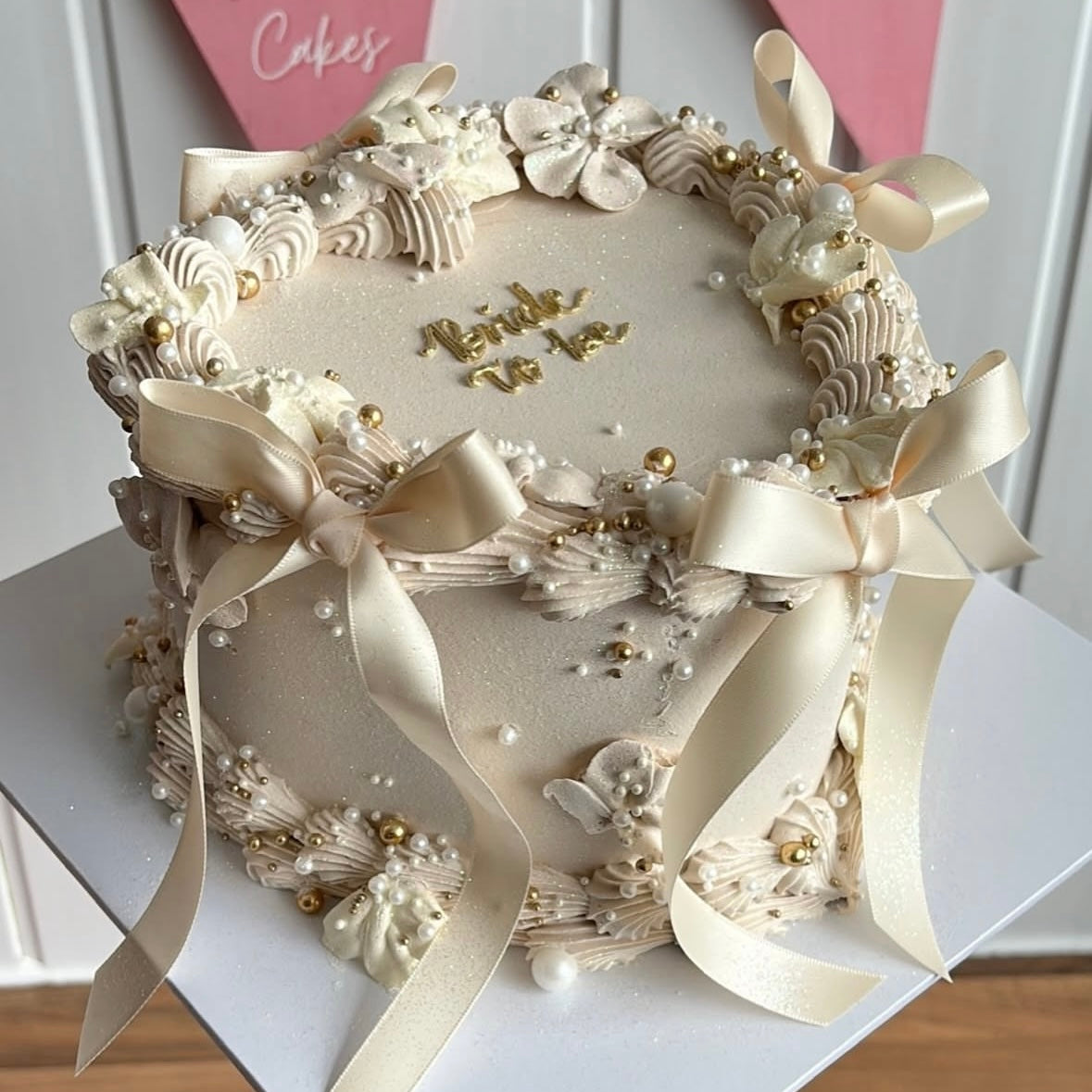 Heart-shaped cake in soft beige, topped with piped buttercream blossoms, delicate ivory satin bows, and scattered gold and white sugar pearls.