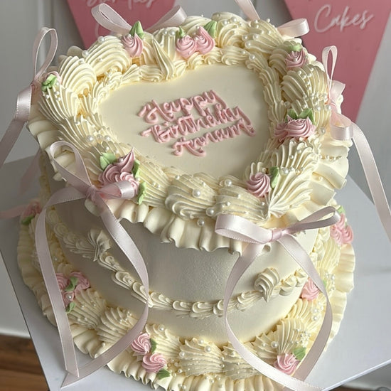 A white/cream buttercream piped vintage style heart cake with pale pink buttercream blossoms, pale pink satin bows and a piped message