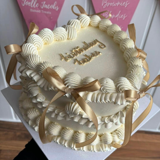 A heart shaped birthday cake with white, piped buttercream frosting in an ornate vintage style. Decorated with shell-like swirls and golden satin ribbon bows at the corners. The cake top has a subtle shimmer and gold lettering that reads “Birthday Babes.”
